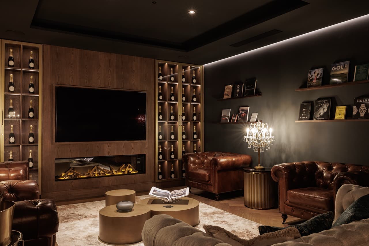 The Clubhouse Marbella - Chesterfield armchairs facing a wood-panelled hearth and screen, illuminated champagne wall on either side, low tungsten light.