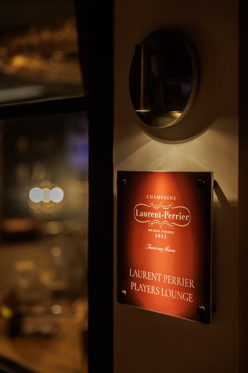 A red-metallic Laurent-Perrier plaque mounted by the entrance to the Players’ Lounge at the Clubhouse Marbella, “Champagne Laurent-Perrier · Maison Fondée 1812 · Tours-sur-Marne · Laurent Perrier Players Lounge” engraved beneath a brass sconce.