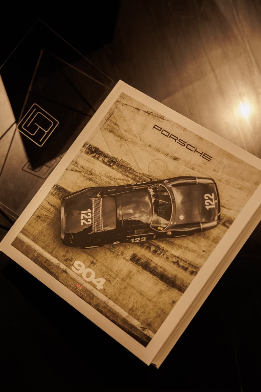 A Porsche 904 monograph resting on a low table at the Clubhouse Marbella, an Olymp Cars-marked display stand to the side under a single warm spotlight.