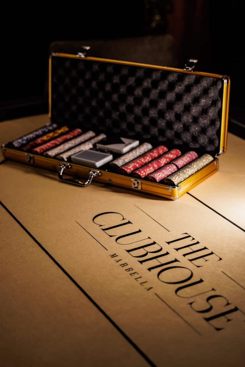 A poker chip case open on a kraft-paper mat printed “The Clubhouse · Marbella”, stacks of chips beside a sealed deck of cards, low tungsten light.