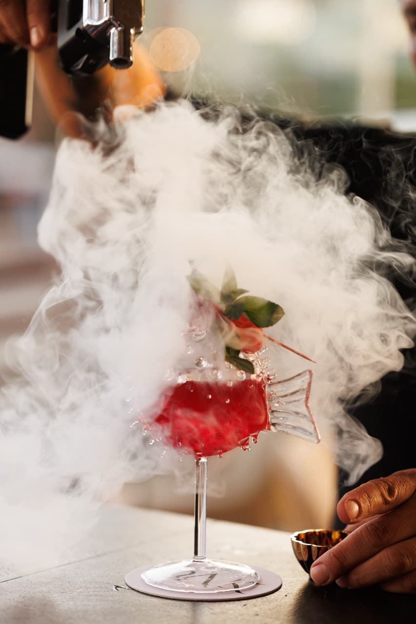 A signature cocktail at La Sala Puerto Banús — a long-stemmed fish-shaped coupe of red liqueur, a strawberry on the rim, dry-ice smoke from a torch above curling around the glass.