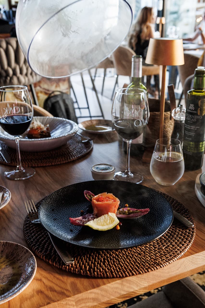 A plate of beef carpaccio at La Sala — thinly sliced cured beef under microgreens on a black ceramic dish, croquetas and olive-oil tasting plates around it, a glass of red wine at the table edge.