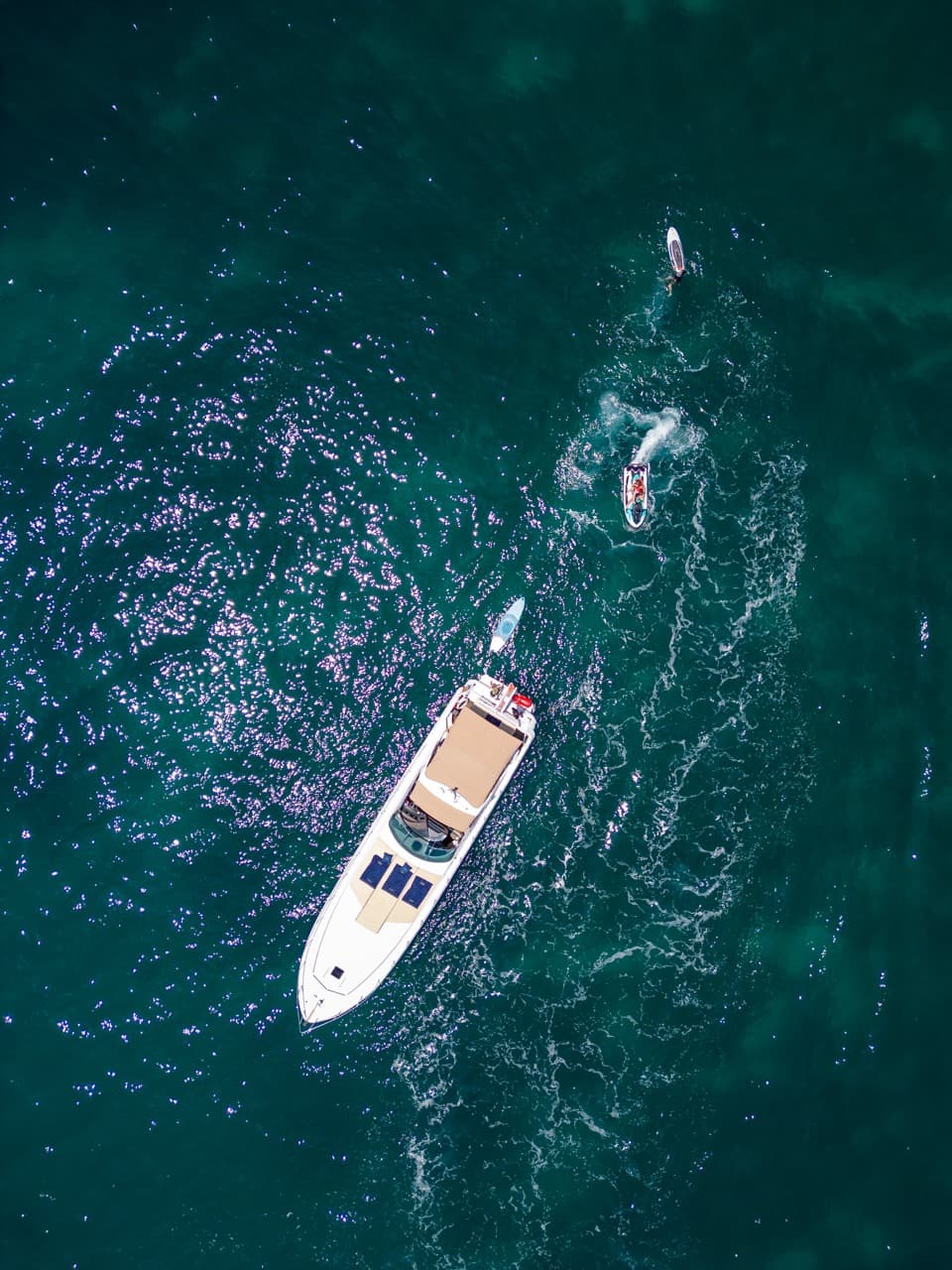 Aerial view of a private yacht at the edge of Puerto Banús with two jet skis trailing white wakes alongside, the surface marbled with reflected light.